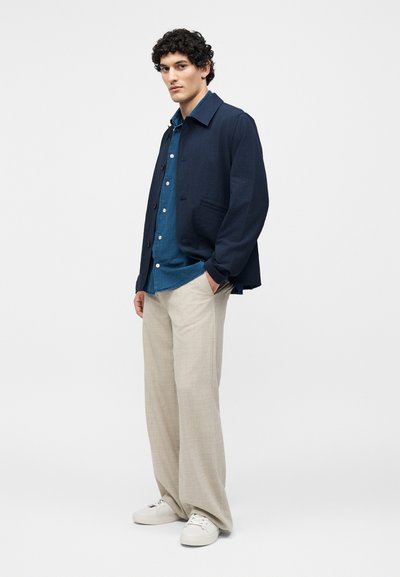 Άνδρας που φοράει ναυτικό μπουφάν πάνω από μπλε πουκάμισο με μπεζ φαρδιά παντελόνια και λευκά αθλητικά παπούτσια, στέκεται με το ένα χέρι στην τσέπη.