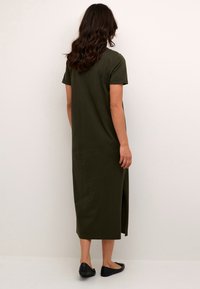 Oliivivihreä polven pituinen mekko, jossa on lyhyet hihat, suora leikkaus, sivu halkioita ja sileä pinta, yhdistettynä mustiin tasapohjaisiin kenkiin.