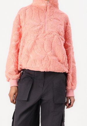 Person trägt einen rosa texturierten Fleecepullover mit halbem Reißverschluss und schwarze Cargohose mit großen Fronttaschen.