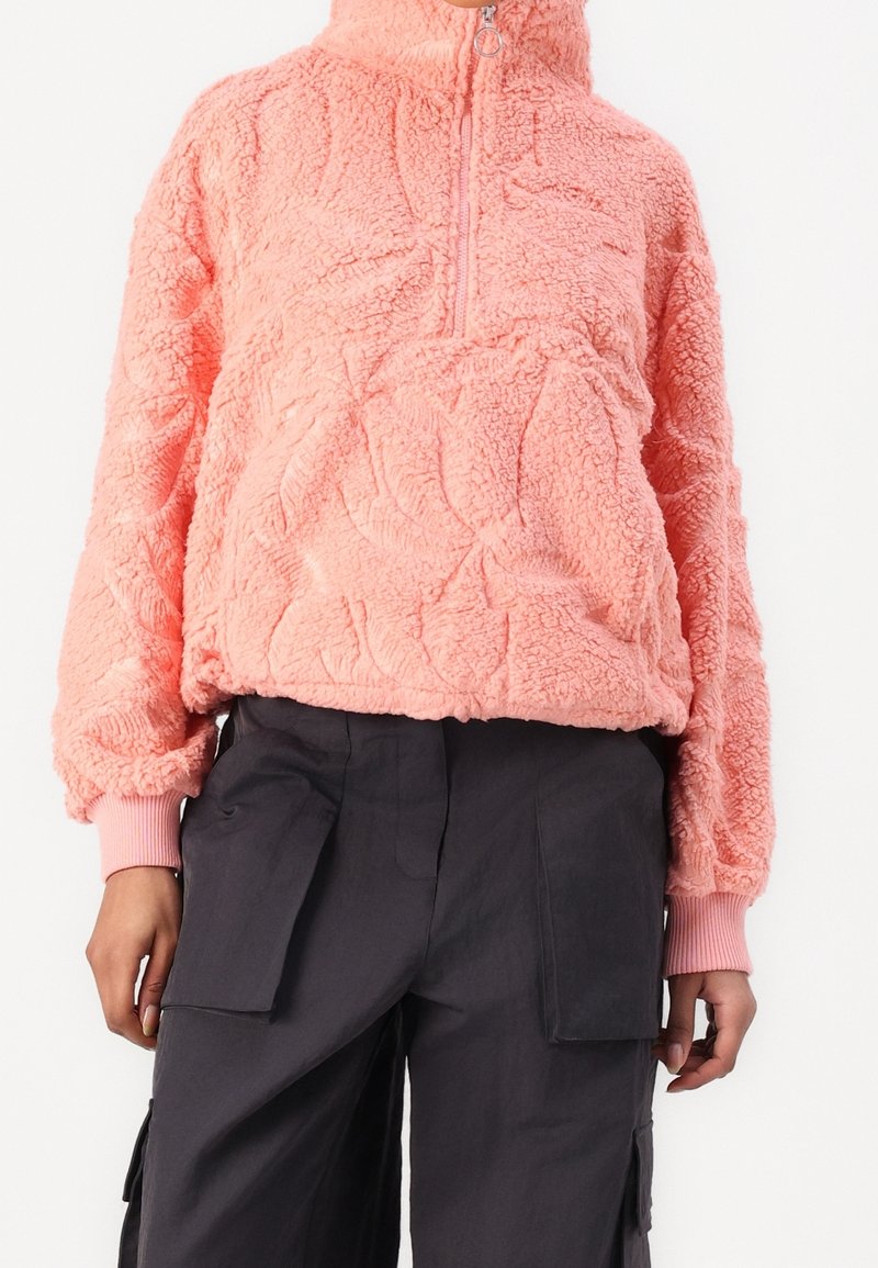 Personne portant un pull en polaire rose texturé avec une fermeture éclair à mi-hauteur et un pantalon cargo noir avec de grandes poches avant.