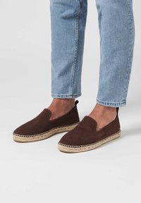 Des chaussures à enfiler en suède marron avec une semelle en jute tressé, présentant un bout rond et des accents élastiques sur les côtés. Portées avec un pantalon en denim bleu clair.