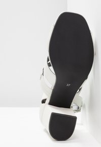 Sandale blanche avec talon bloc, semelle noire visible, taille 37, inclinée contre un fond blanc d’angle.
