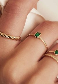 Goldene Ringe an einer Hand, mit einem gedrehten Band, einem rechteckigen grünen Stein und einem Band, das mit kleinen klaren Steinen akzentuiert ist. Glatte Textur.