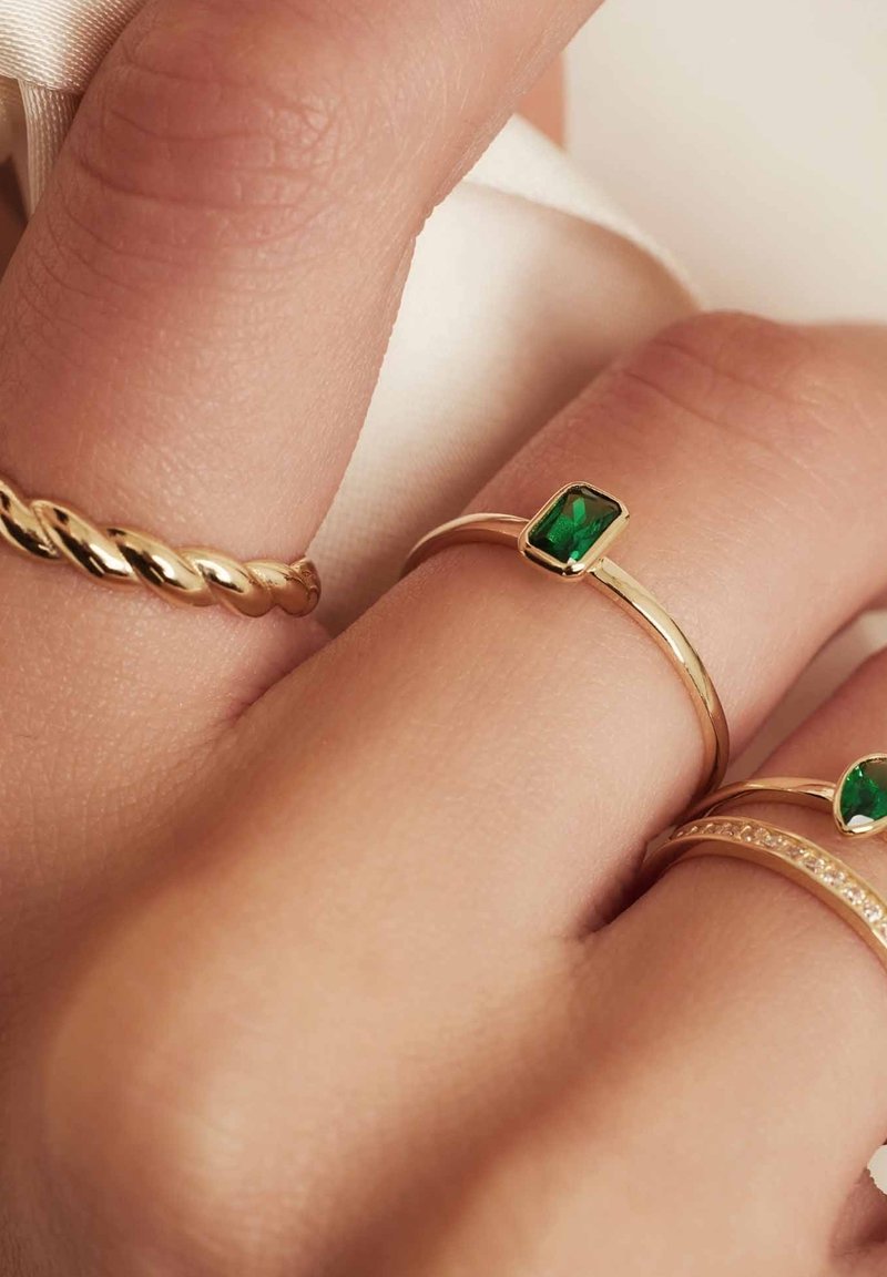 Goldene Ringe an einer Hand, mit einem gedrehten Band, einem rechteckigen grünen Stein und einem Band, das mit kleinen klaren Steinen akzentuiert ist. Glatte Textur.