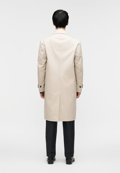 Μπεζ μακρύ παλτό με ευθεία γραμμή, μονό πίσω άνοιγμα, ορατά κουμπιά και λεία υφή, συνδυασμένο με σκούρα παντελόνια με λεπτές ρίγες και παπούτσια.