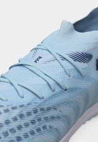 Scarpa da ginnastica azzurro chiaro con tomaia in maglia, lacci integrati e dettagli scuri a motivi sul puntale e sul lato.