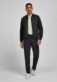 Herren Bomberjacke mit einer strukturierten schwarzen Vorderseite und marineblauen Ärmeln, kombiniert mit dunklen Hosen und weißen Turnschuhen. Schlichtes, modernes Design.