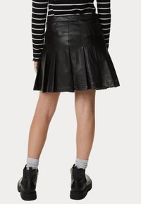 Falda de cuero negra con pliegues y costuras visibles. Llevada con botas negras hasta el tobillo y calcetines grises, mostrada desde la parte de atrás.