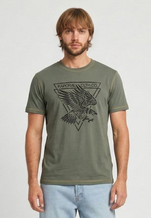 Homme portant un t-shirt vert olive avec un motif d'aigle noir et le texte « KAPORAL CLTH.CO. », associé à un jean bleu clair.