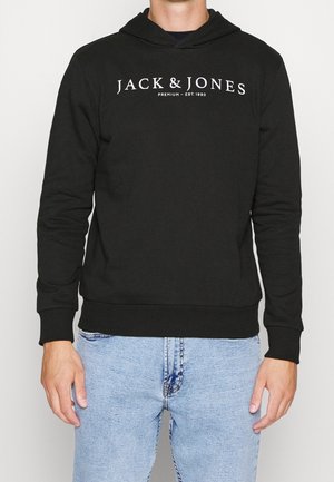 Persona che indossa una felpa nera con cappuccio con il testo "JACK & JONES PREMIUM – EST. 1990" e jeans azzurri, in piedi contro uno sfondo chiaro e uniforme.