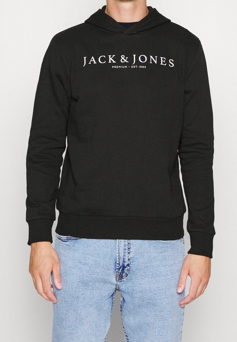Personne portant un sweat à capuche noir avec le texte "JACK & JONES PREMIUM – EST. 1990" et un jean bleu clair, debout devant un fond clair uni.