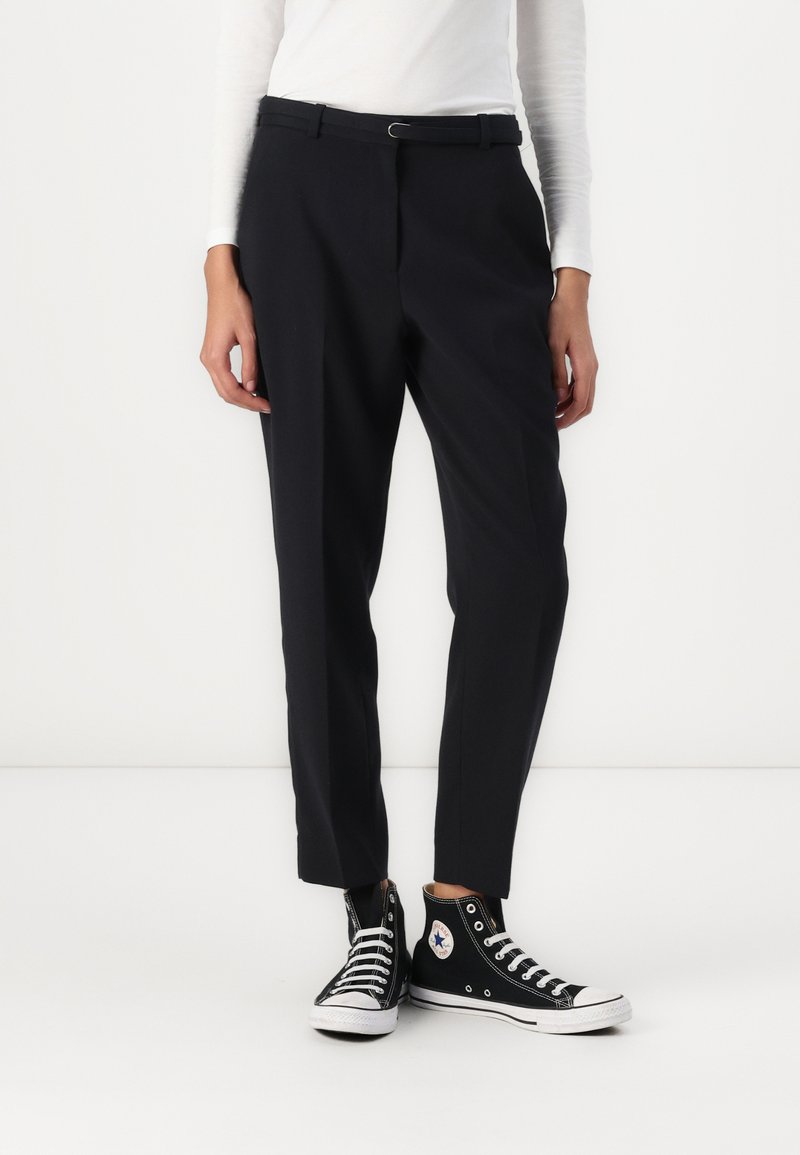 Pantalons noirs ajustés avec une coupe slim, présentant une taille avec passants de ceinture et une texture lisse, associés à des baskets montantes noires.