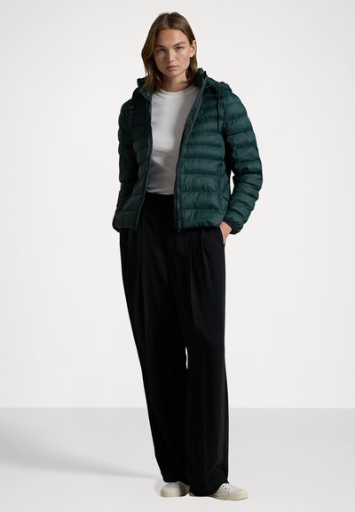 Donkergroene gewatteerde jas met capuchon, ritsluiting, horizontaal gequilt patroon, gedragen over een wit shirt en zwarte broek, gecombineerd met witte schoenen.