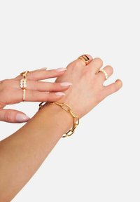 Smycken i guldton med ett kraftigt länkarmband, flera ringar med olika designer och långa, spetiga naglar. Släta och glänsande ytor.