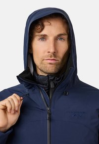 Giacca blu navy con cappuccio, dotata di zip frontale, corde regolabili e logo ricamato "SWIDEMOUNT". Indossata sopra uno strato interno texturizzato.