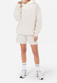 Sweat à capuche crème avec une poche avant et un logo, associé à un short assorti. La tenue est composée d'un tissu léger et de manches longues, portée avec des baskets blanches.