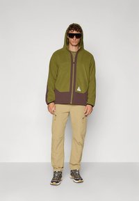 Zelena in rjava flis jakna s kapuco, z zadrgo po celotni dolžini, sprednjimi žepi in trikotnim logotipom. Kombinirana s bež cargo hlačami in pohodniškimi čevlji.