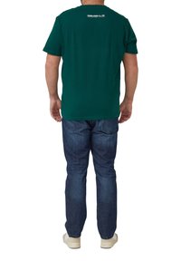 Mann trägt ein grünes kurzärmliges T-Shirt und blaue Jeans, steht mit entspannten Armen und dem Rücken zum Betrachter vor einem weißen Hintergrund.