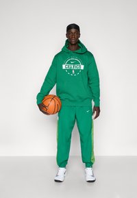 Grøn Nike hoodie med "CELTICS" logo, parret med grønne Nike sweatpants med sidestriber, holder en basketball, hvid sneakers.