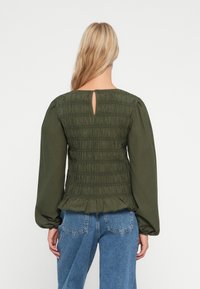 Blouse vert olive avec un dos smocké, un ourlet volanté et des manches longues. Présente une ouverture en forme de clé à l'arrière et une texture de tissu lisse.