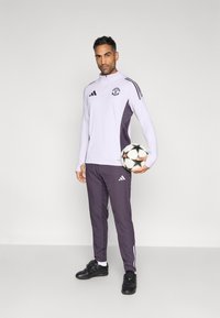 Λευκή και μωβ φόρμα Adidas με ζακέτα φερμουάρ και αντίστοιχο παντελόνι, με λογότυπο, μαύρα παπούτσια, κρατώντας μια μπάλα ποδοσφαίρου με αστέρια.