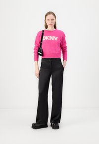 Vaaleanpunainen pörröinen neule, jossa on "DKNY" -logo, yhdistettynä korkeavyötäröisiin mustiin housuihin ja massiivisiin mustiin platform-kengiin. Yksinkertainen musta olkalaukku.