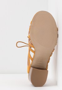 Yellow high-heeled sandal with crisscross strap design, smooth leather upper, textured rubber sole, and embossed brand details on the sole.