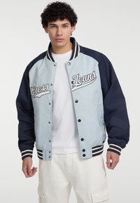 Lichtblauwe en marineblauwe bomberjack met geborduurd "Guess Jeans" logo. Heeft knoppen met drukknoopsluiting, gestreepte manchetten en een soepele textuur.