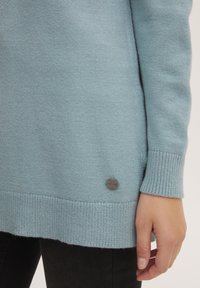 Svetlo modri pulover iz mehkega materiala z rebrastimi manšetami in robom. Ima majhen okrogli kovinski detajl blizu dna, na levi strani.