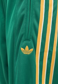 Πράσινο αθλητικό παντελόνι από ύφασμα με κίτρινες και λευκές ρίγες και κίτρινο κεντημένο λογότυπο κοντά στην τσέπη με φερμουάρ.