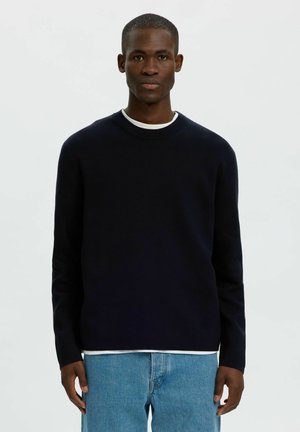 Jeune homme noir portant un pull noir à manches longues sur une chemise blanche et un jean bleu clair, debout devant un fond clair uni.
