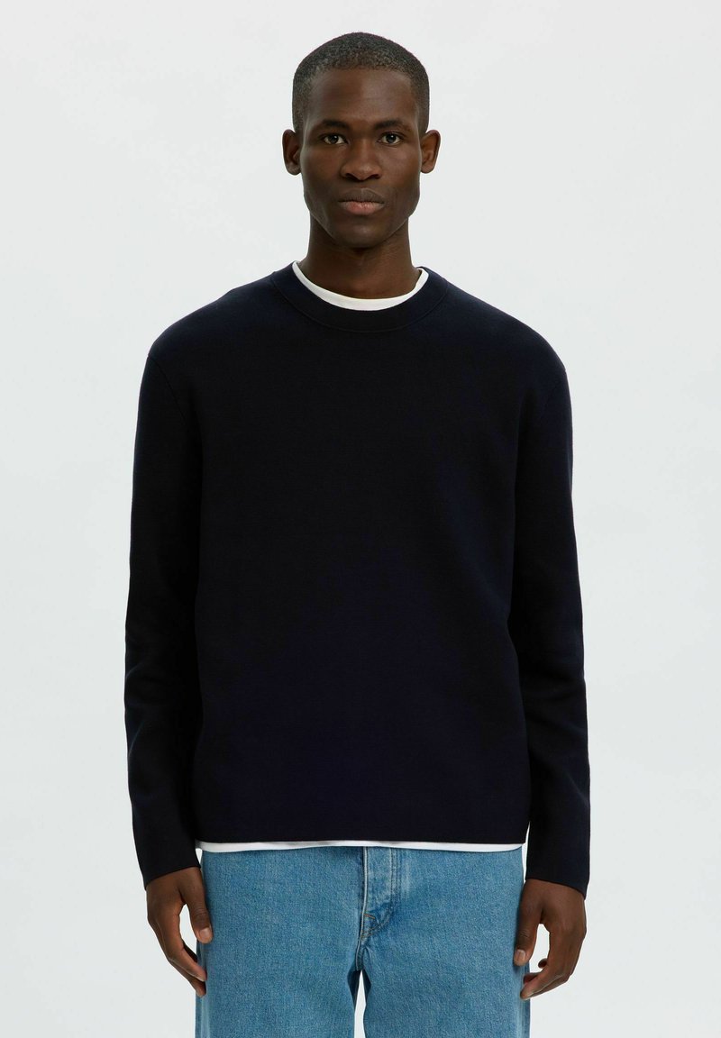 Jeune homme noir portant un pull noir à manches longues sur une chemise blanche et un jean bleu clair, debout devant un fond clair uni.