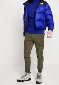 Modrá péřová bunda s černými doplňky, modré tričko s dlouhým rukávem, olivové cargo kalhoty a šedé turistické boty. Má elastické manžety a kapsy na zip.