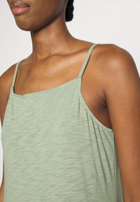 Débardeur vert clair avec des fines bretelles, présentant un tissu doux et texturé et un design à coupe décontractée, s'étendant légèrement aux épaules.