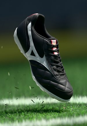 Zwart-witte voetbalschoen in de lucht boven gras op een voetbalveld met een witte lijn op de achtergrond.