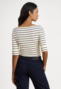 Γυναίκα με μακριά σκούρα μαλλιά φοράει λευκό πουκάμισο με λεπτές οριζόντιες μπλε ρίγες και μπλε παντελόνι, στέκεται πλευρικά με τα χέρια στις τσέπες.