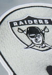 Schwarz-weiß gesticktes Logo mit einem Totenschädel, der eine Augenklappe trägt, darunter gekreuzte Schwerter, in einem schildförmigen Umriss.
