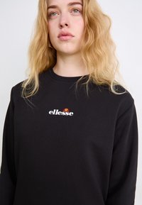 Sudadera negra con cuello redondo y mangas largas. Presenta un logo blanco de "ellesse" con acentos rojos y naranjas. Textura de tejido suave.