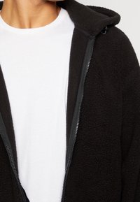 Nahaufnahme einer Person, die ein weißes T-Shirt mit Rundhalsausschnitt unter einer offenen schwarzen Fleecejacke trägt, die einen sichtbaren Reißverschluss und Kragen hat.