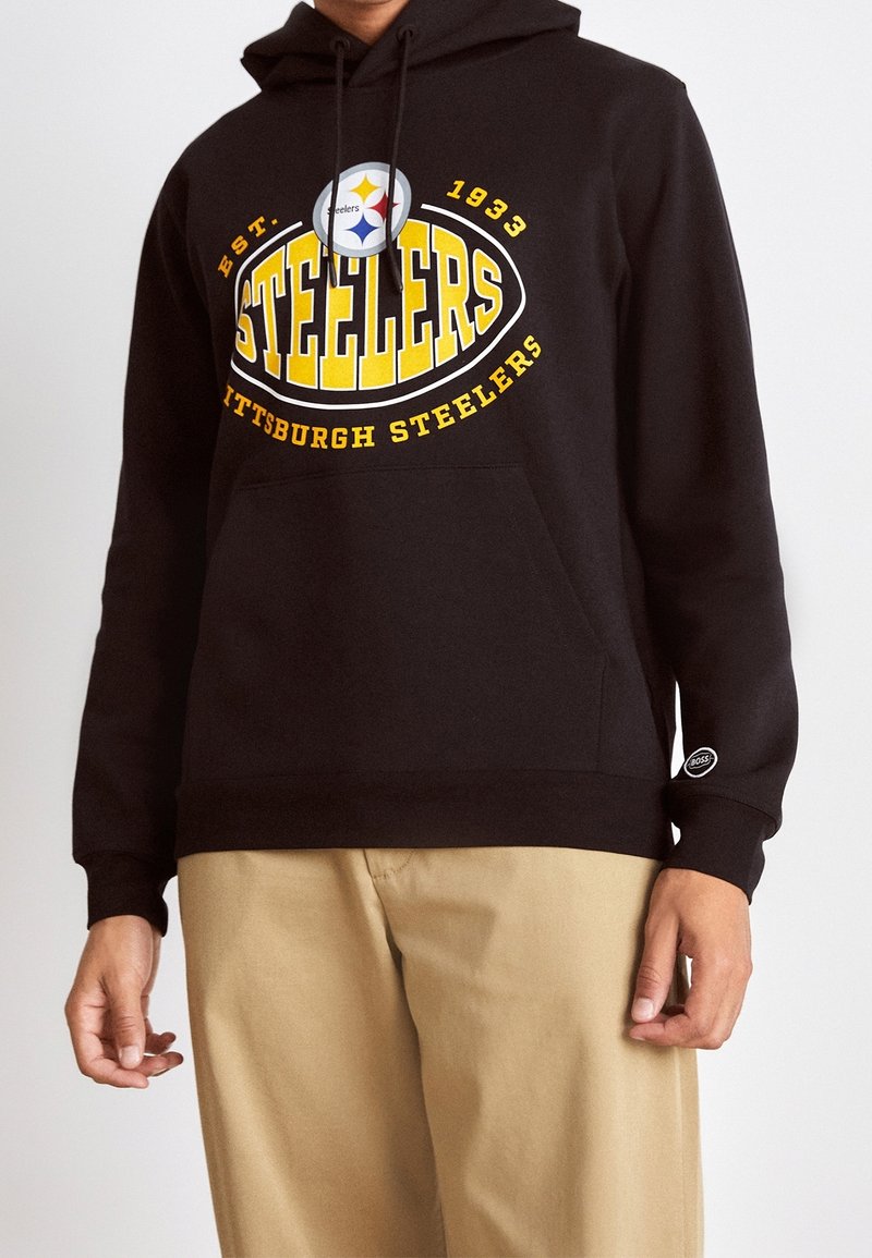 Personne portant un sweat à capuche noir des Pittsburgh Steelers avec un texte et un logo jaunes, associé à un pantalon beige clair.