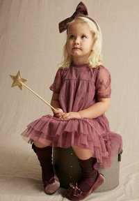 Lila Tüllkleid mit gestickten Details, geschichteter Rüschensockel, kombiniert mit dunklen Socken und weichen Schuhen. Das Kind hält einen goldenen Sternenstab.