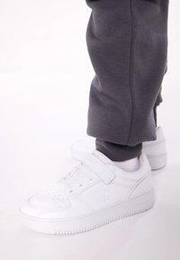 Baskets blanches en cuir lisse, dotées d'une sangle Velcro et d'un détail de logo discret, accompagnées de pantalons de survêtement gris.