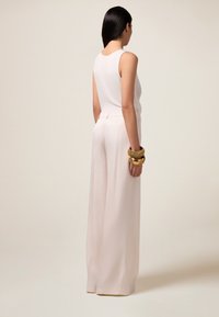 Lichtroze jumpsuit met een ronde halslijn en een wijde pijpen-silhouet. De stof lijkt glad. Het model draagt gouden armbanden om één pols.