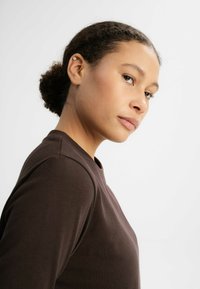 Braunes Langarmshirt mit rundem Ausschnitt, das sich durch eine glatte Textur und eine enganliegende Form auszeichnet, vor einem schlichten hellen Hintergrund präsentiert.