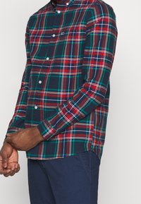 Camisa de cuadros abotonada con patrones en azul marino, rojo, verde y amarillo; mangas largas; dobladillo redondeado; y cuello abotonado.