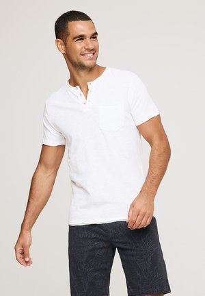 Hombre sonriente con una camisa henley blanca de manga corta con botones y pantalones cortos azul marino con estampado de hojas, de pie frente a un fondo liso.