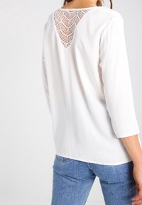 Vit blus med trekvartslånga ärmar och en triangulär spetsdetalj på baksidan. Slät tyg och avslappnad passform. Denimjeans synliga nederst.