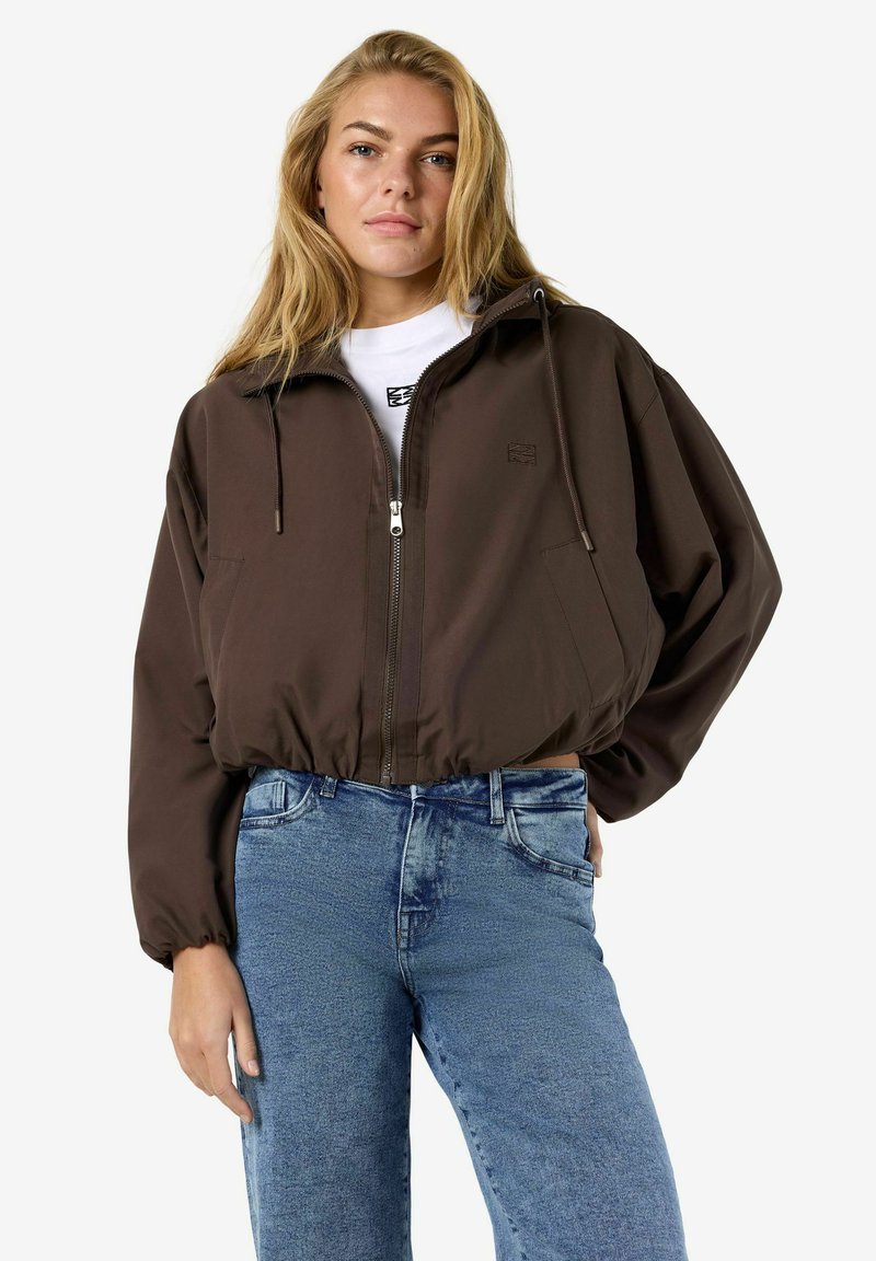 Jeune femme aux longs cheveux blonds portant une veste marron à zip et un jean bleu clair, debout avec une main sur la hanche.