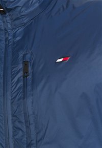 Cazadora azul marino con cierre de cremallera, superficie texturizada y un pequeño logo con detalles en rojo, blanco y azul en el pecho.