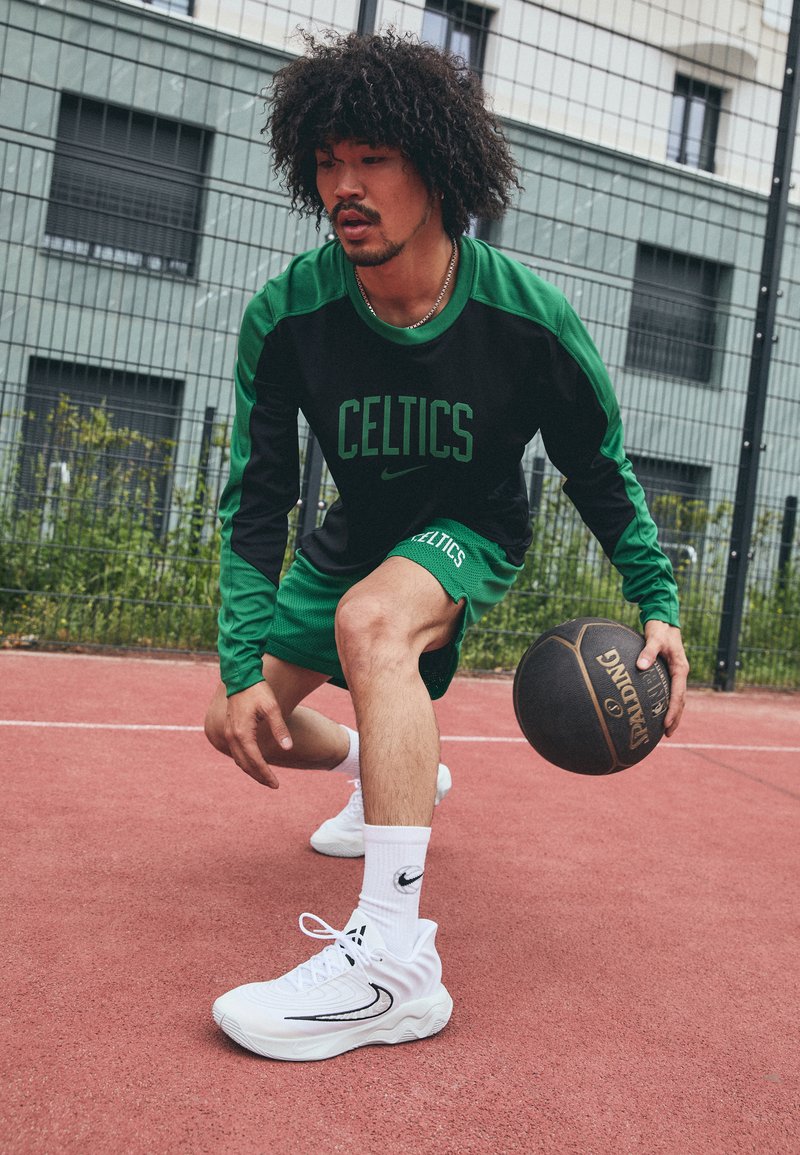 Ropa deportiva verde y negra con el texto "CELTICS", zapatillas blancas y balón de baloncesto negro en una cancha roja. Tejido texturizado y ajuste atlético.
